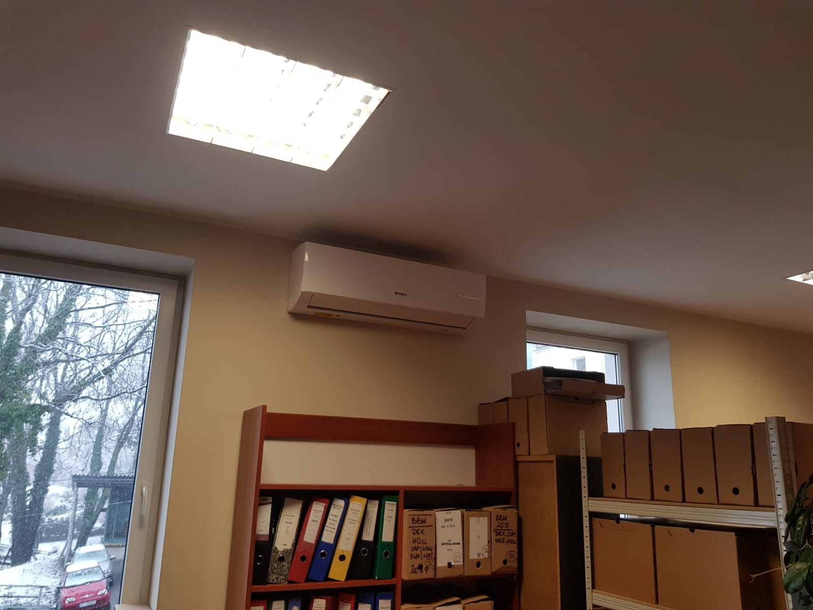 Biały klimatyzator ścienny marki Gree zamontowany w biurze nad regałem z segregatorami i kartonami, widok z dołu na tle okna z widokiem na zimowe drzewa.