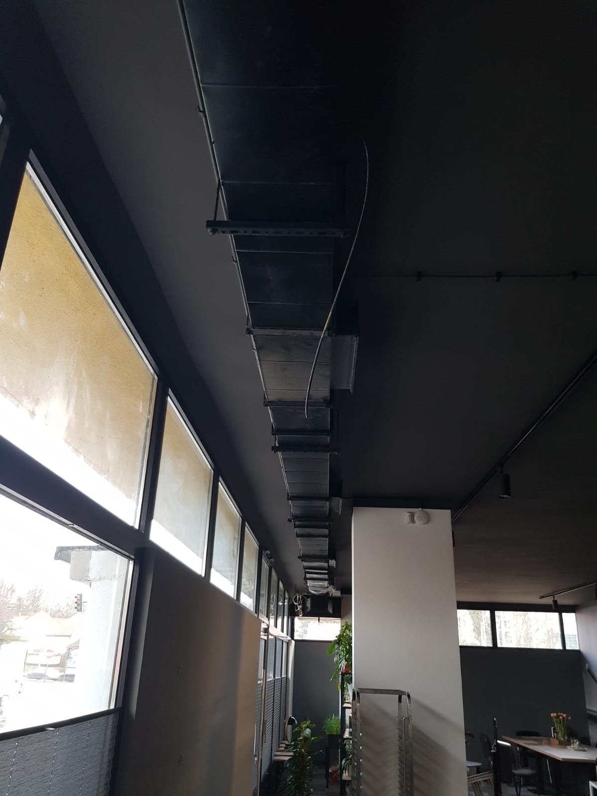 Szereg przewodów wentylacyjnych prostokątnych, zawieszonych pod czarnym sufitem w budynku komercyjnym, widoczne okna z roletami i rośliny doniczkowe.