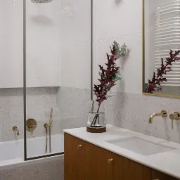 Nowoczesna łazienka z białymi, pionowo ułożonymi płytkami nad wanną, szarym lastryko na ścianach i podłodze, oraz drewnianą szafką umywalkową z złotymi detalami, uzupełniona szklanym wazonem...