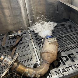 Warsztat Wolica - Czyszczenie filtra DPF w specjalnej myjni. Widoczne zanieczyszczenia wypływające z filtra. Metalowa konstrukcja myjni i elementy mocujące filtr.