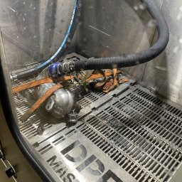 Warsztat Wolica - Filtr DPF w komorze czyszczącej DPF Master, zabezpieczony pasami. Widoczny wąż doprowadzający płyn czyszczący. Metalowa kratka na dnie komory.