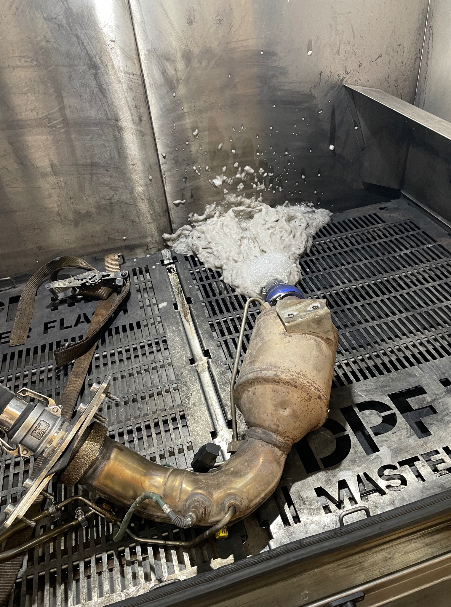 Czyszczenie filtra DPF w specjalnej myjni. Widoczne zanieczyszczenia wypływające z filtra. Metalowa konstrukcja myjni i elementy mocujące filtr.