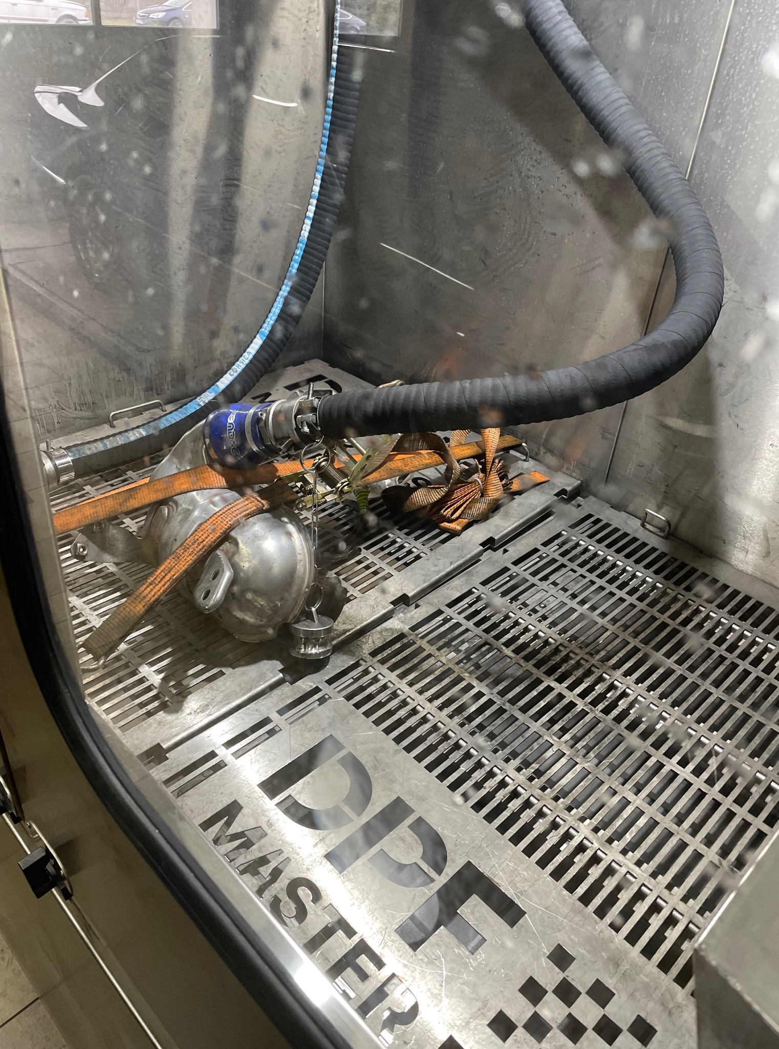 Filtr DPF w komorze czyszczącej DPF Master, zabezpieczony pasami. Widoczny wąż doprowadzający płyn czyszczący. Metalowa kratka na dnie komory.