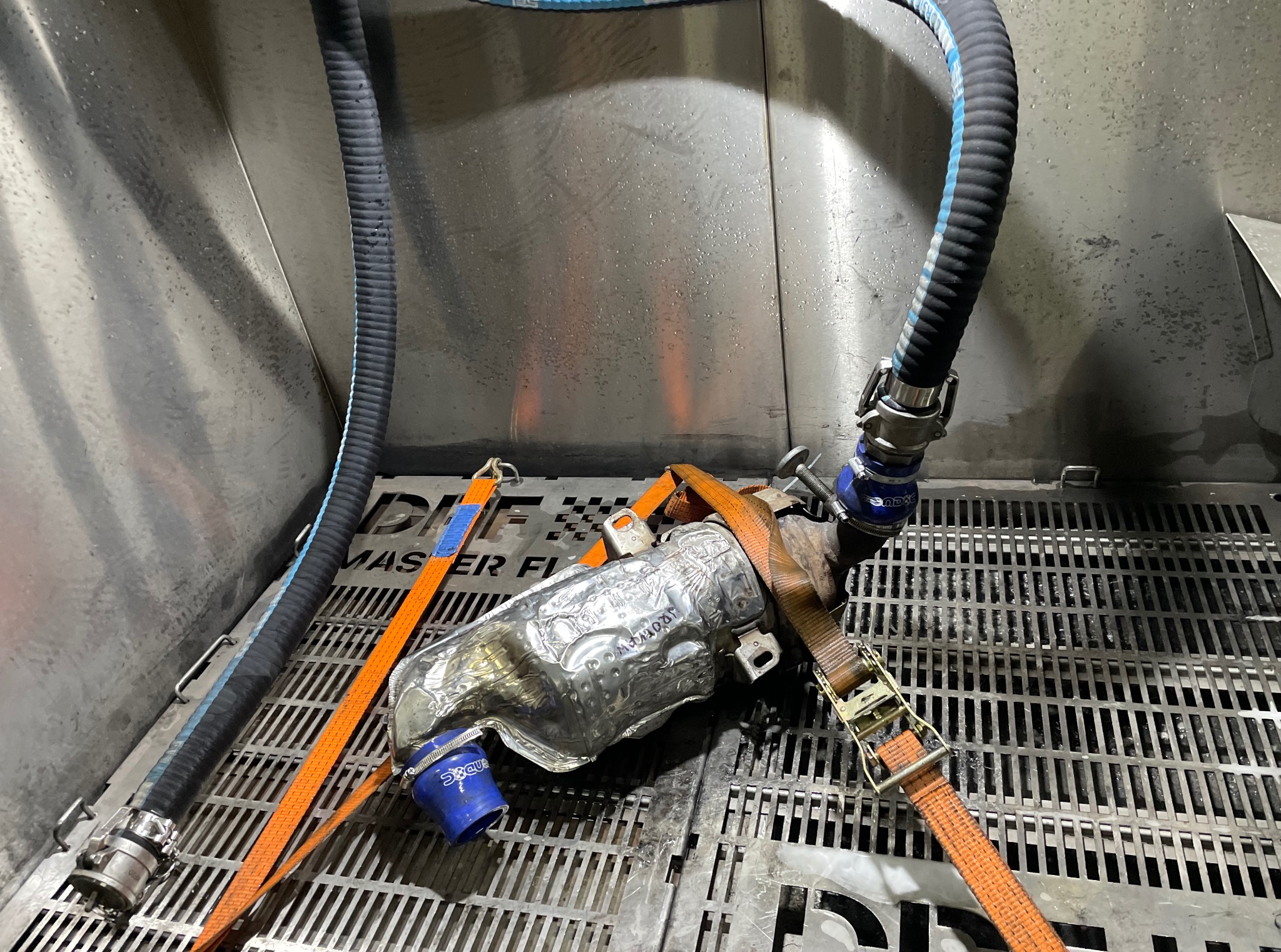 Filtr cząstek stałych (DPF) w metalowej komorze, zabezpieczony pomarańczowymi pasami, z podłączonymi wężami. Widoczne ślady czyszczenia.