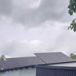 Panele słoneczne zamontowane na dachu budynku gospodarczego w pochmurny dzień, widoczne z dołu, z fragmentami drzew w kadrze.