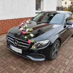 Czarny Mercedes udekorowany kwiatami i tablicą rejestracyjną 'Nowożeńcy' stoi na brukowanym podjeździe.