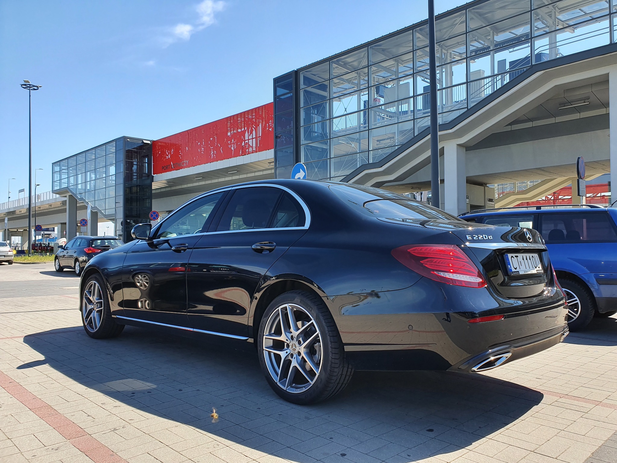 Czarny samochód osobowy Mercedes E220d zaparkowany przed nowoczesnym budynkiem dworca z przeszkloną fasadą i czerwonymi akcentami.