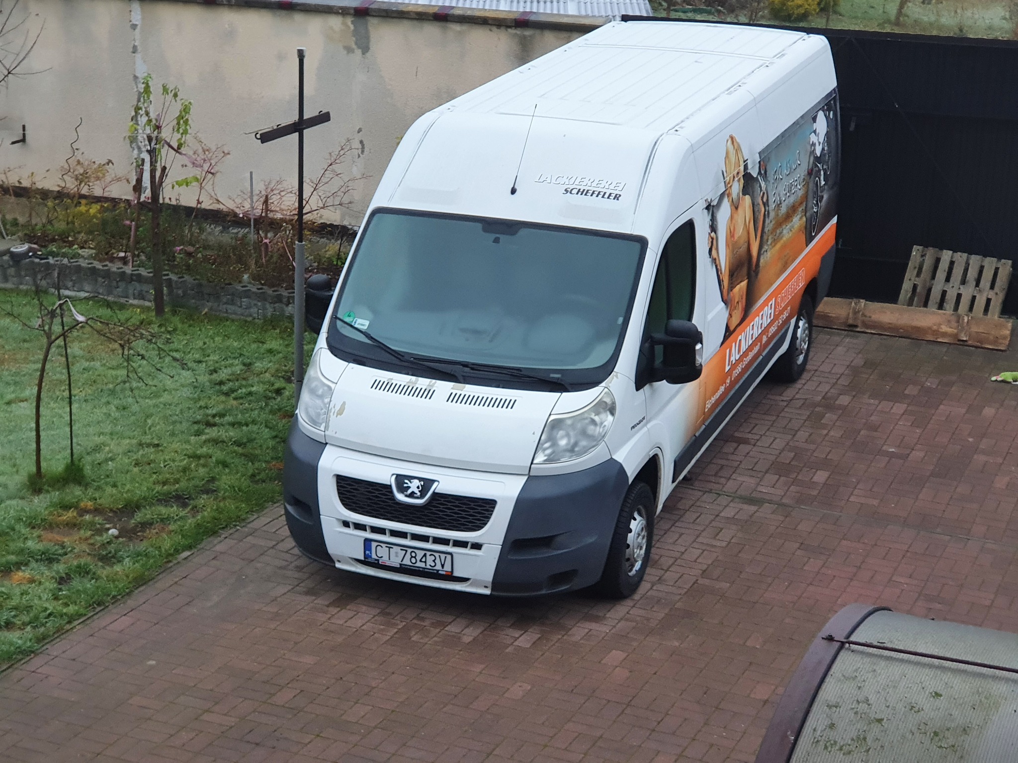 Biały samochód dostawczy z grafiką i napisem 'Lackiererei Scheffler' zaparkowany na brukowanym podjeździe, widok z góry.