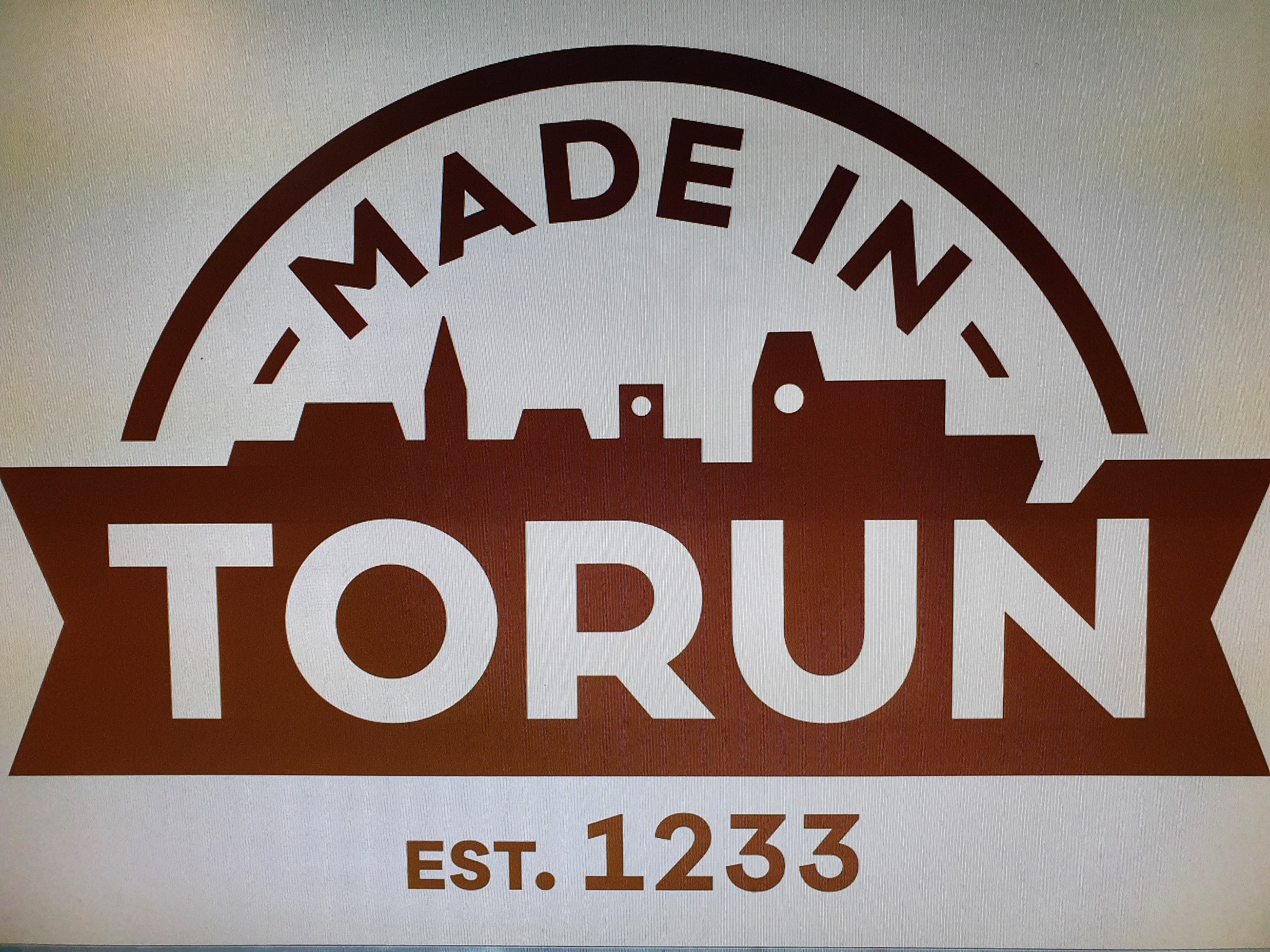 Brązowo-białe logo z napisem 'MADE IN' w łuku nad grafiką sylwetki miasta, poniżej napis 'TORUŃ' w prostokątnym banerze, a pod nim 'EST. 1233'.