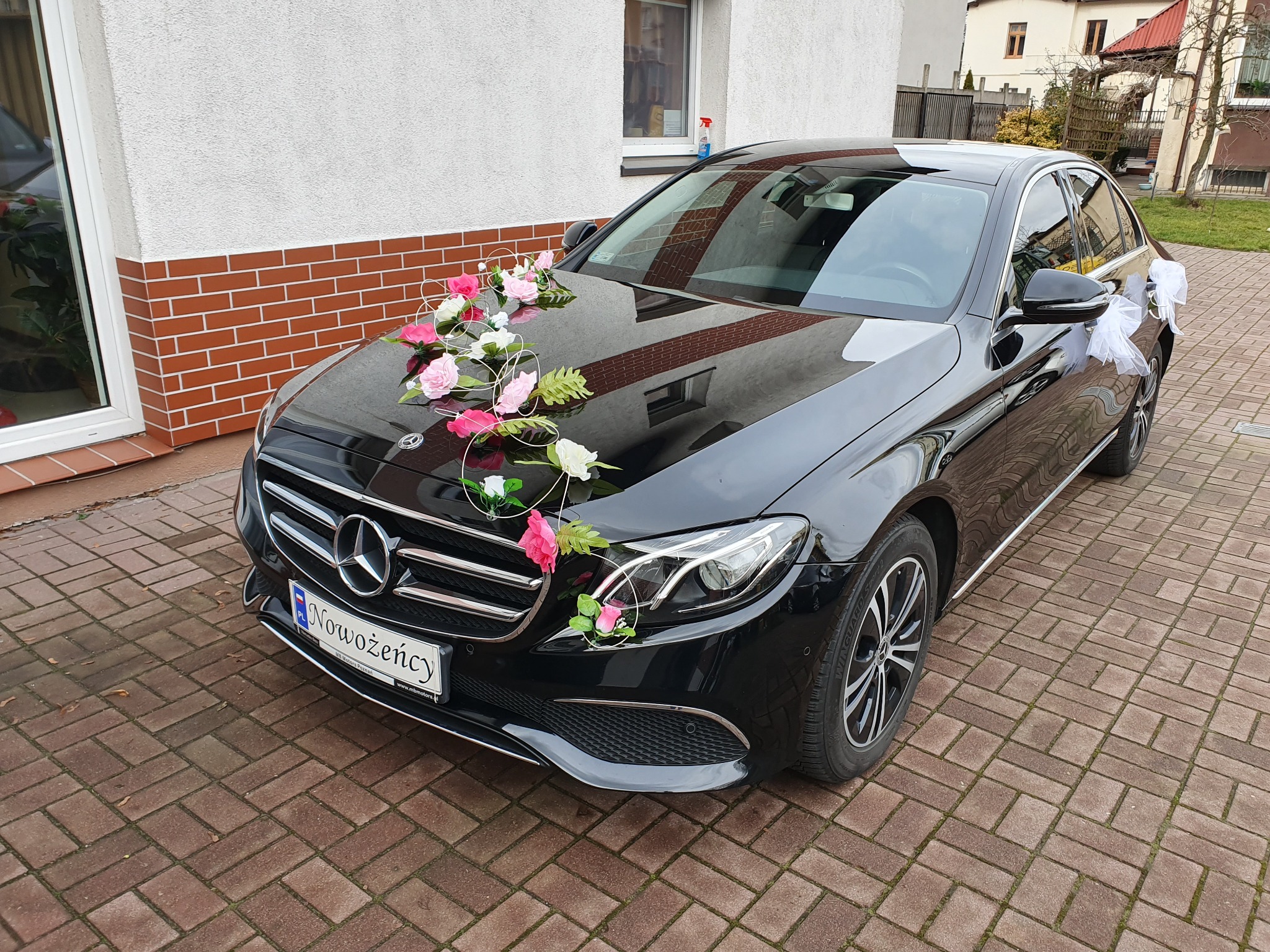 Czarny Mercedes udekorowany kwiatami i tablicą rejestracyjną 'Nowożeńcy' stoi na brukowanym podjeździe.