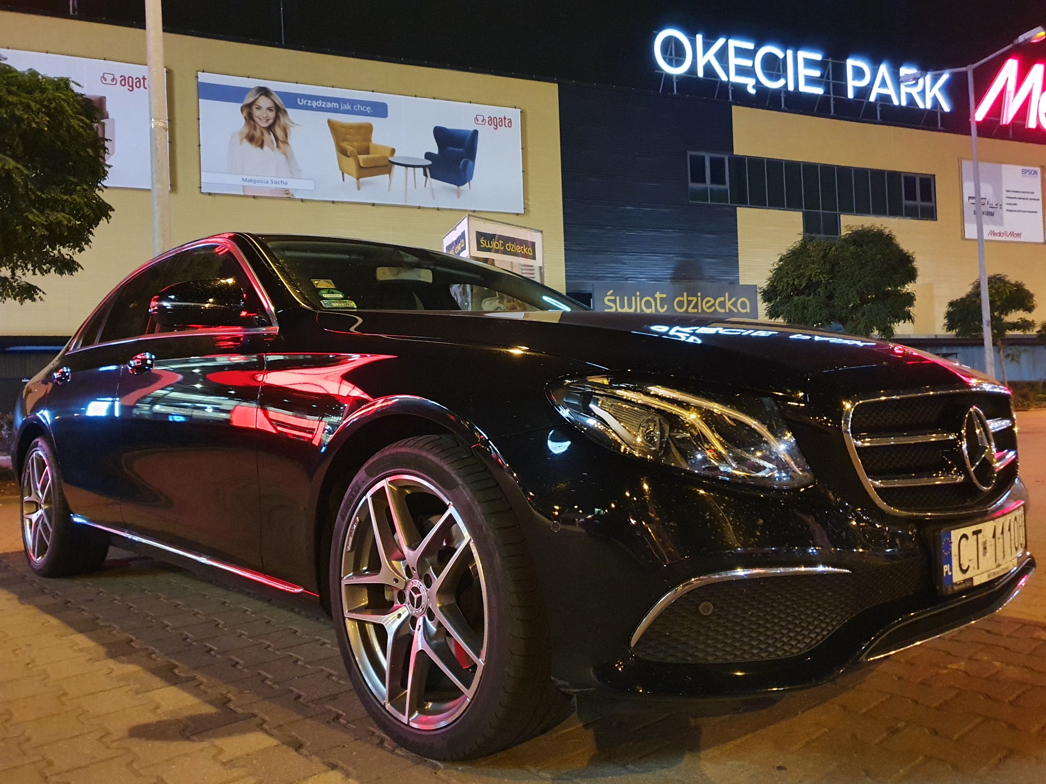 Czarny Mercedes-Benz E-klasa zaparkowany nocą przed centrum handlowym Okęcie Park, z widocznym numerem rejestracyjnym i odbiciami świateł w karoserii.