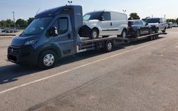 Laweta z trzema samochodami osobowymi i dostawczymi, widok z boku, szary ciągnik, jasne tło parkingu.