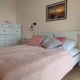 Jasna sypialnia z białymi meblami: komoda, łóżko z różową narzutą i poduszkami, szafka nocna, lampy ścienne z dekoracyjnymi żarówkami, obraz z motywem plaży nad łóżkiem.