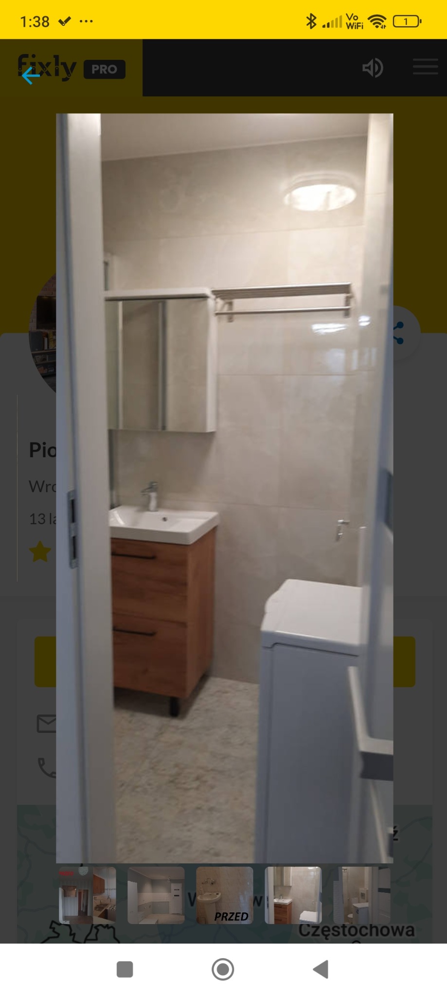 Nowoczesna łazienka z drewnianą szafką pod umywalką, lustrem z szafką nad umywalką, pralką i beżowymi płytkami na ścianach, widziana zza otwartych drzwi.