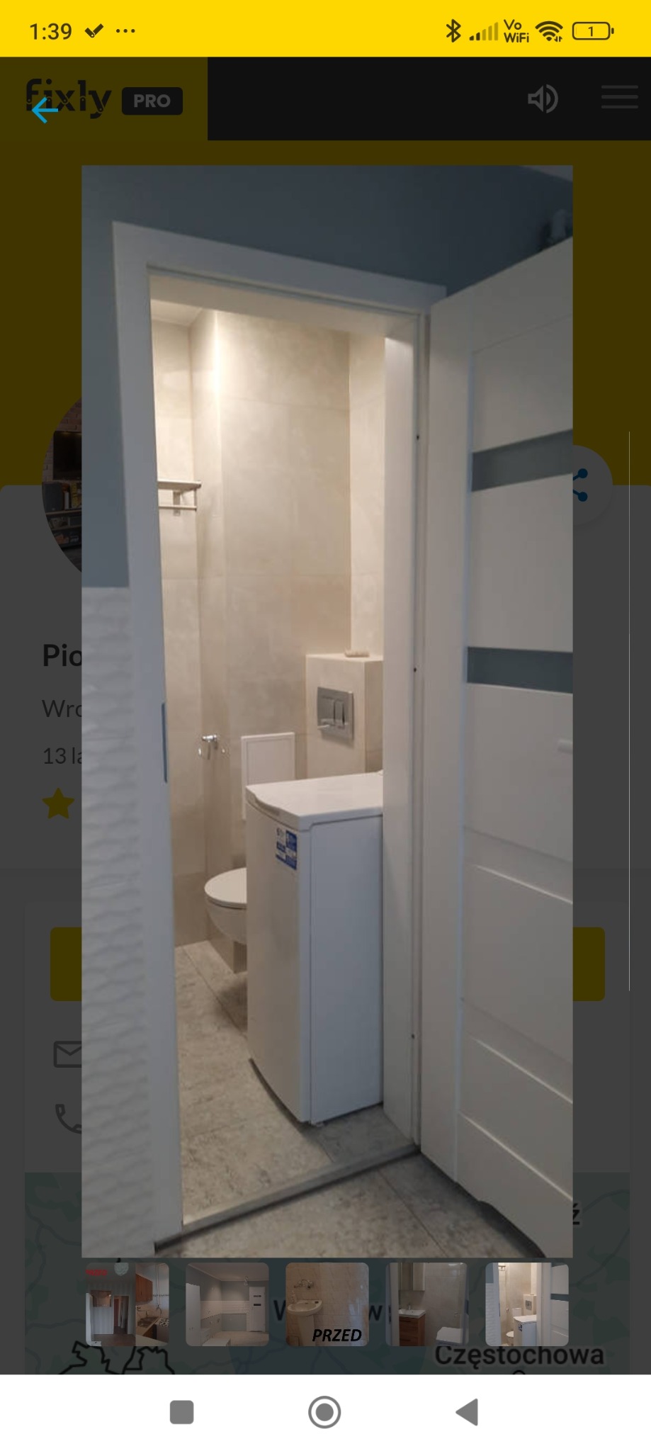 Wąska łazienka z pralką, wykończona w jasnych płytkach, widok zza uchylonych białych drzwi z szarymi wstawkami.