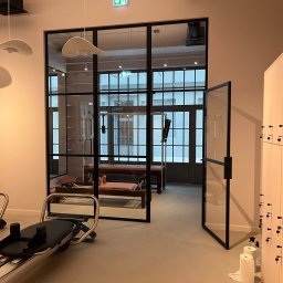 Pro_Poziom - Jasne wnętrze studia pilates z czarną, przeszkloną ścianą działową i otwartymi drzwiami, widoczne maszyny do ćwiczeń i szafki z czarnymi gałkami, na podłodze rolki papieru i butelki ze sprayem.