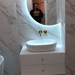 Pro_Poziom - Biała umywalka nablatowa na szafce z frezowanymi frontami i złotymi gałkami, okrągłe lustro z podświetleniem LED, płytki imitujące marmur na ścianach i podłodze, widoczne baterie umywalkowe...