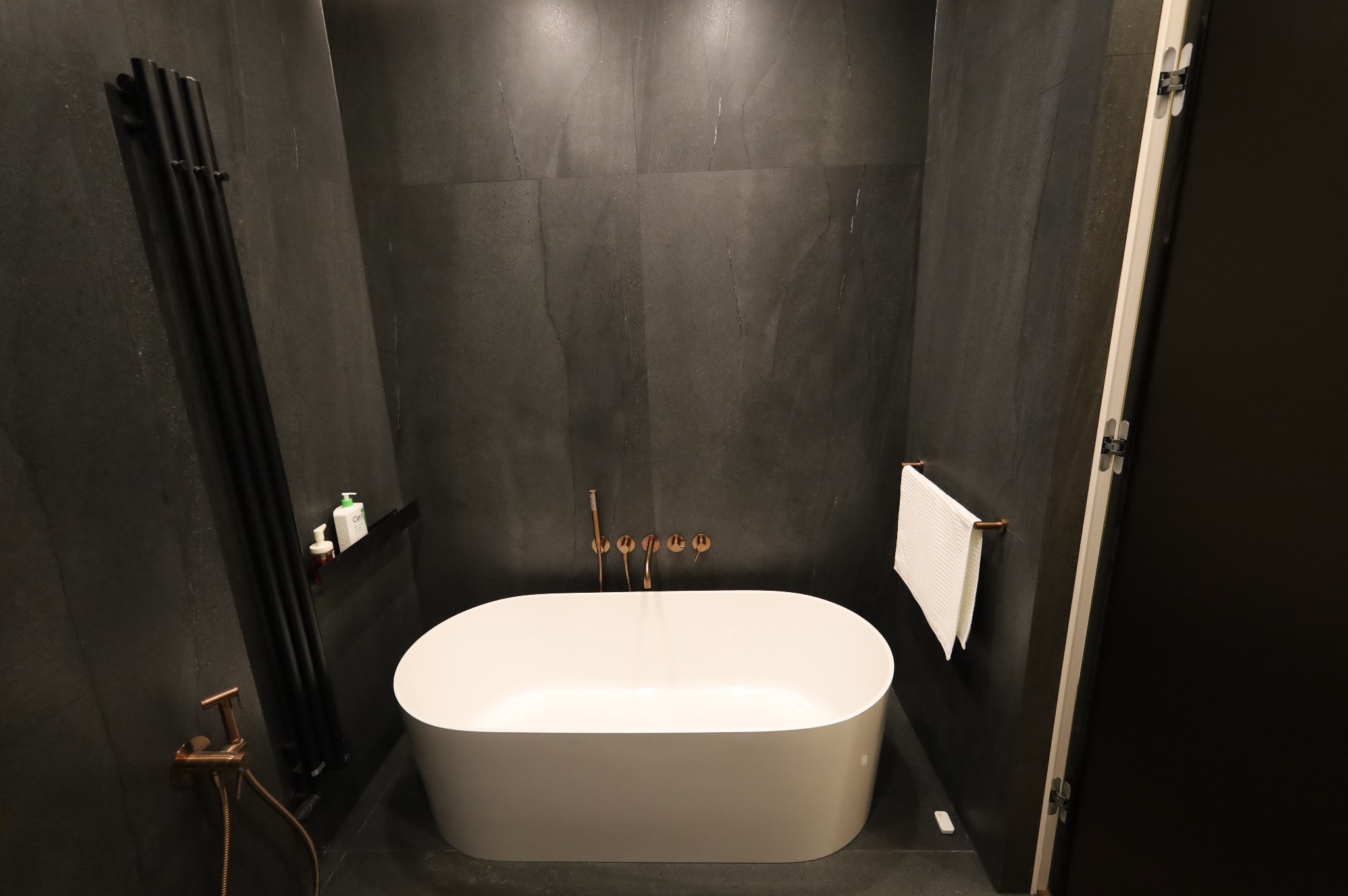 Nowoczesna łazienka z czarnymi płytkami, owalną białą wanną wolnostojącą i miedzianą armaturą ścienną, minimalistyczny grzejnik drabinkowy w kolorze czarnym, biały ręcznik na miedzianym wieszaku.