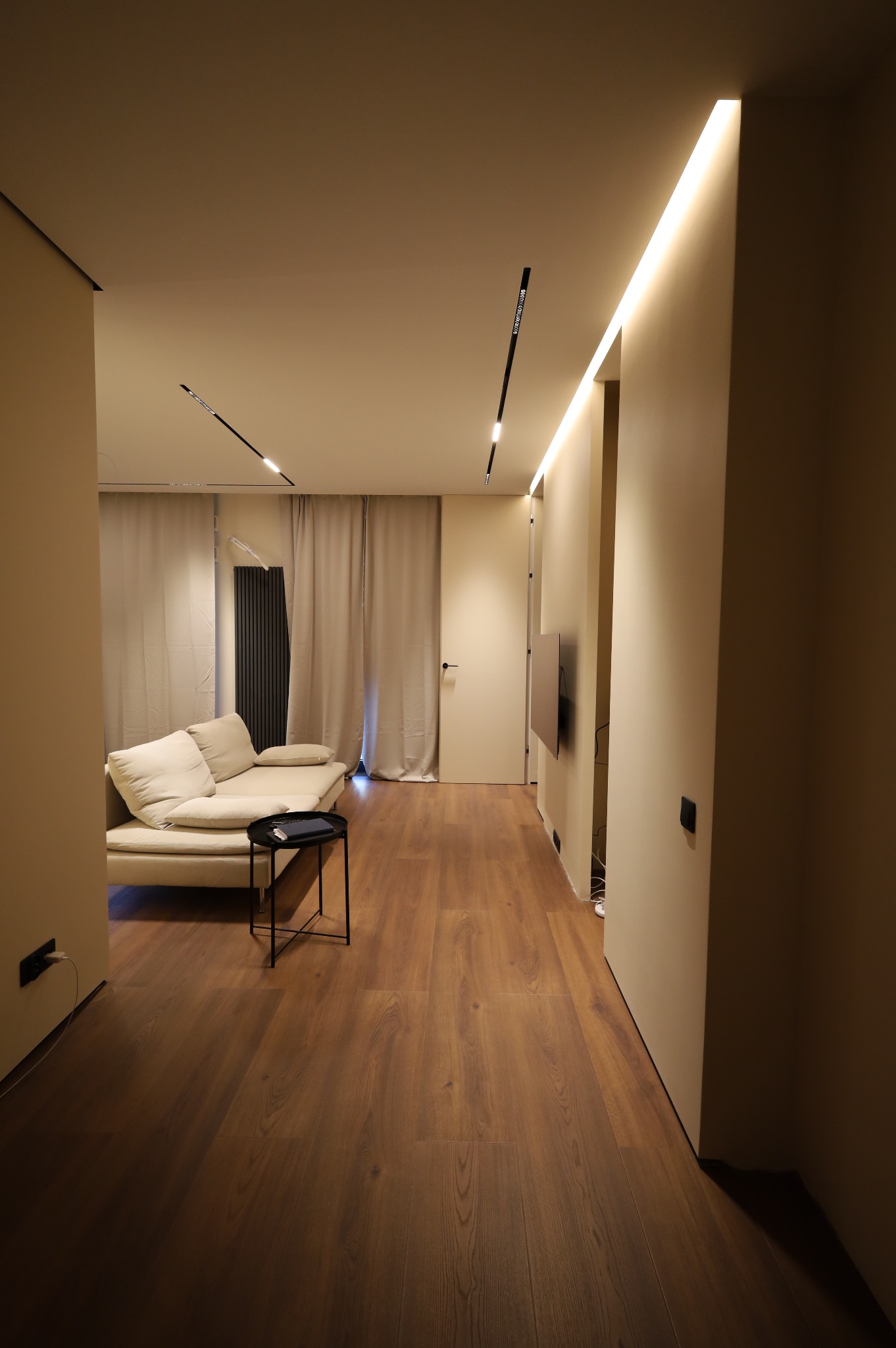 Nowoczesny salon z drewnianą podłogą, beżowymi ścianami, minimalistyczną sofą i liniowym oświetleniem LED w suficie.