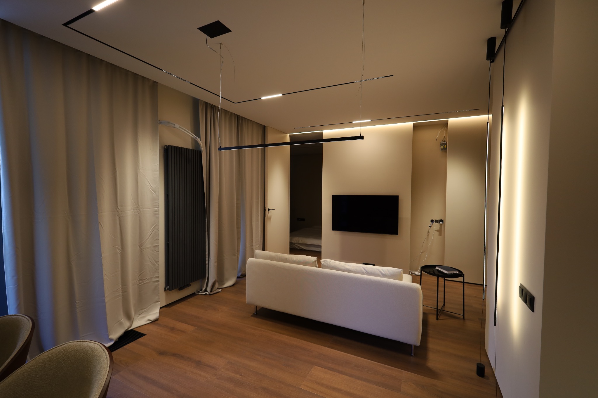 Nowoczesne wnętrze mieszkania z drewnianą podłogą, beżowymi ścianami, minimalistycznym oświetleniem, białym narożnikiem i zasłoniętymi oknami.