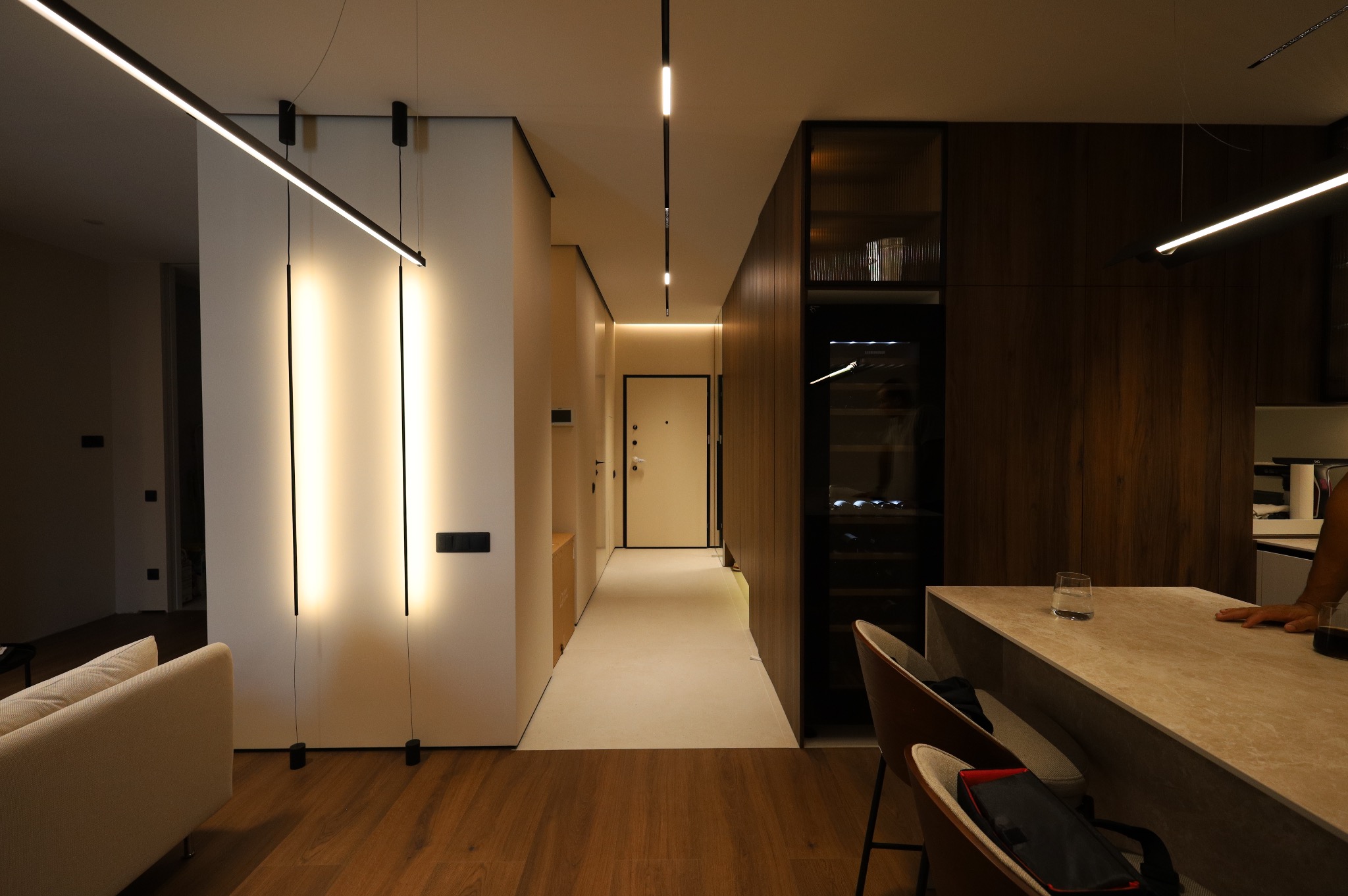 Nowoczesne wnętrze mieszkania z oświetleniem LED: pionowe lampy na ścianie, podłużne lampy sufitowe, drewniana zabudowa z barkiem i chłodziarką do wina, oraz wyspa kuchenna z krzesłami.
