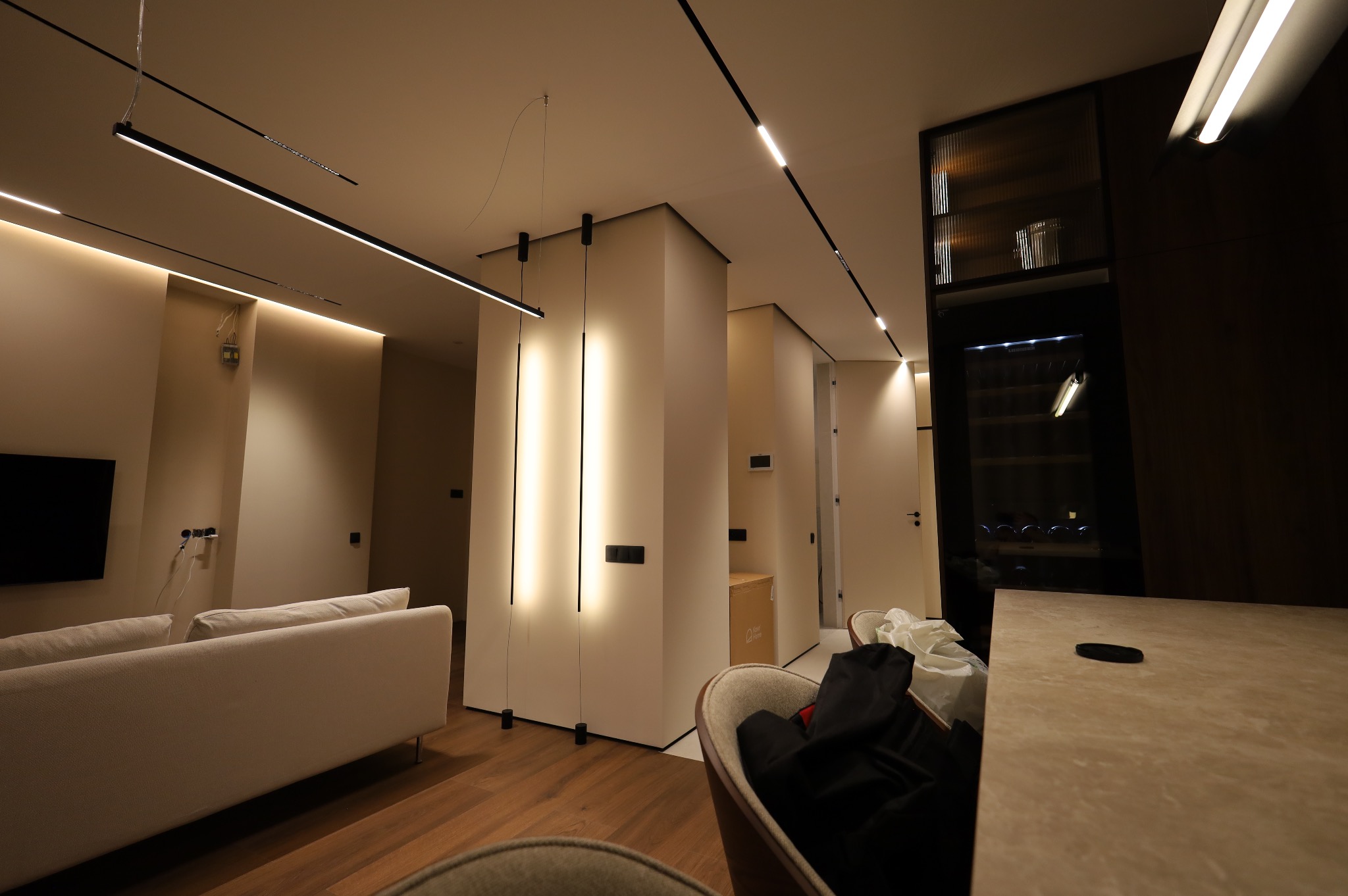 Nowoczesne wnętrze mieszkania z oświetleniem liniowym LED na suficie i pionowymi panelami LED w ścianach, widoczny fragment sofy i drewniana podłoga.
