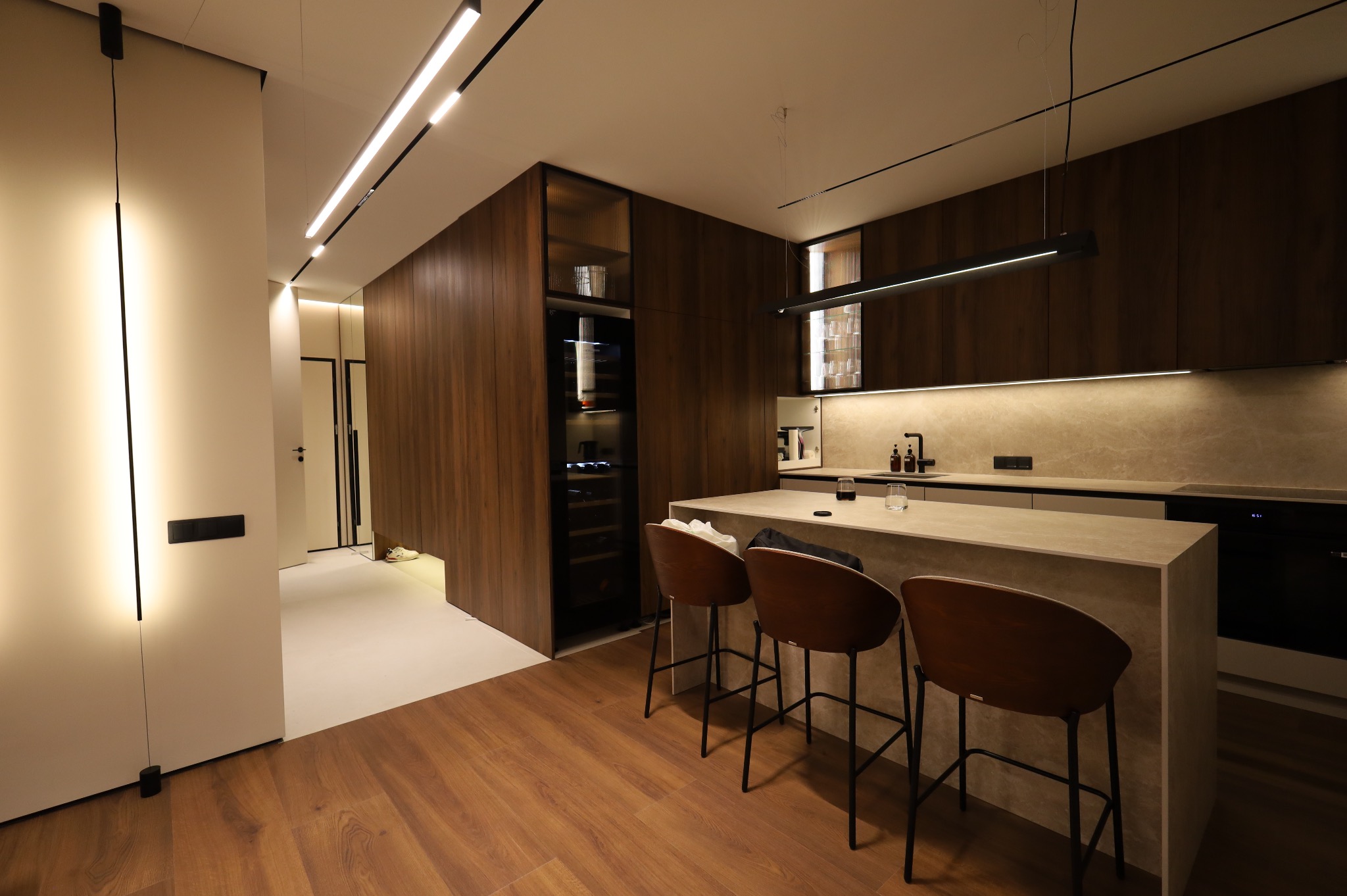 Nowoczesna kuchnia z drewnianymi frontami, wyspą z kamiennym blatem i trzema hokerami, oświetlona listwami LED w suficie i pionową lampą przy ścianie.