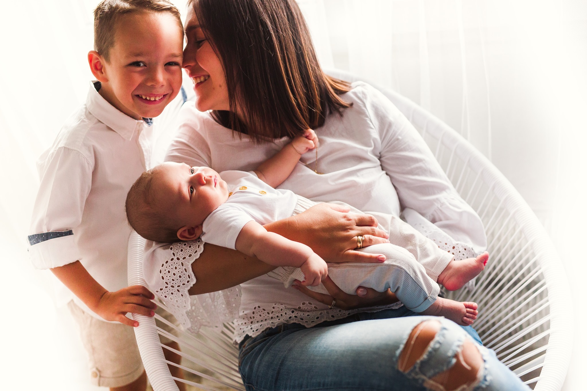 Uśmiechnięta matka siedzi w białym fotelu, trzyma niemowlę, obok stoi starszy syn w białej koszuli, wszyscy patrzą na siebie w jasnym świetle.