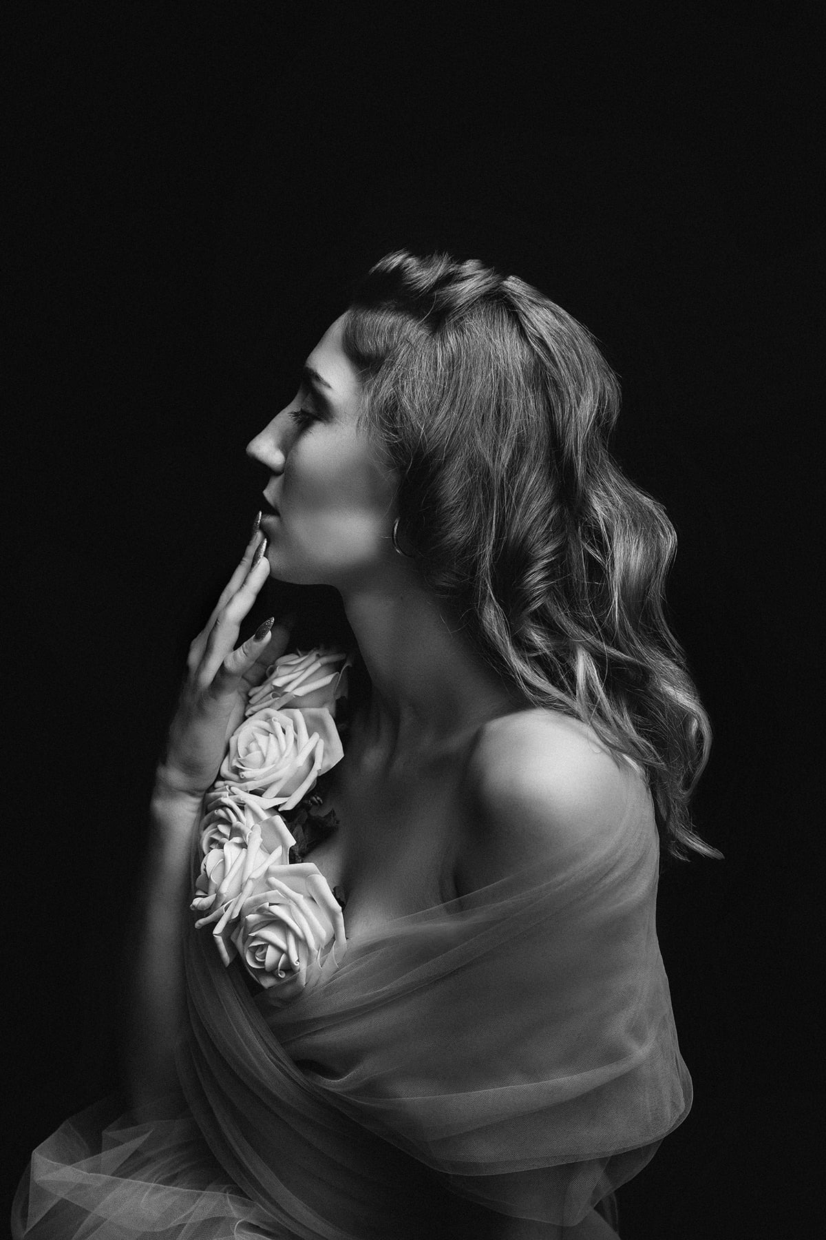 Czarno-biały portret kobiety z profilu, z dłonią przy ustach i białymi różami na ramieniu, owiniętej w delikatny materiał na ciemnym tle.