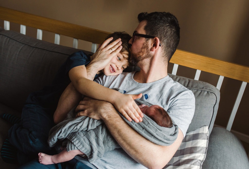 Ojciec w okularach siedzi na szarej kanapie, trzyma noworodka owiniętego w szary koc, starszy syn obejmuje ich oboje i uśmiecha się.