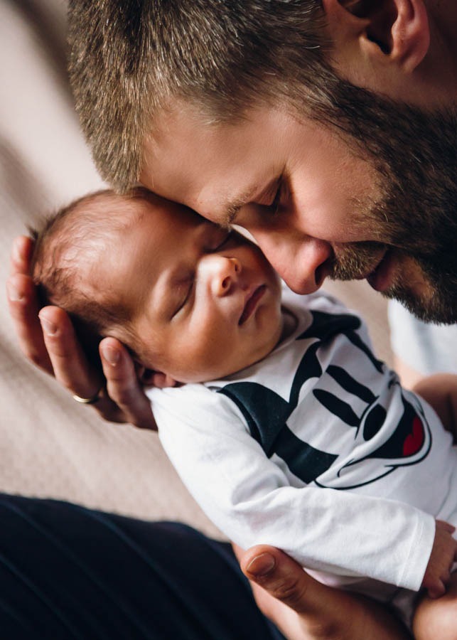 Ojciec z brodą czule przytula śpiącego noworodka ubranego w body z motywem Myszki Miki.