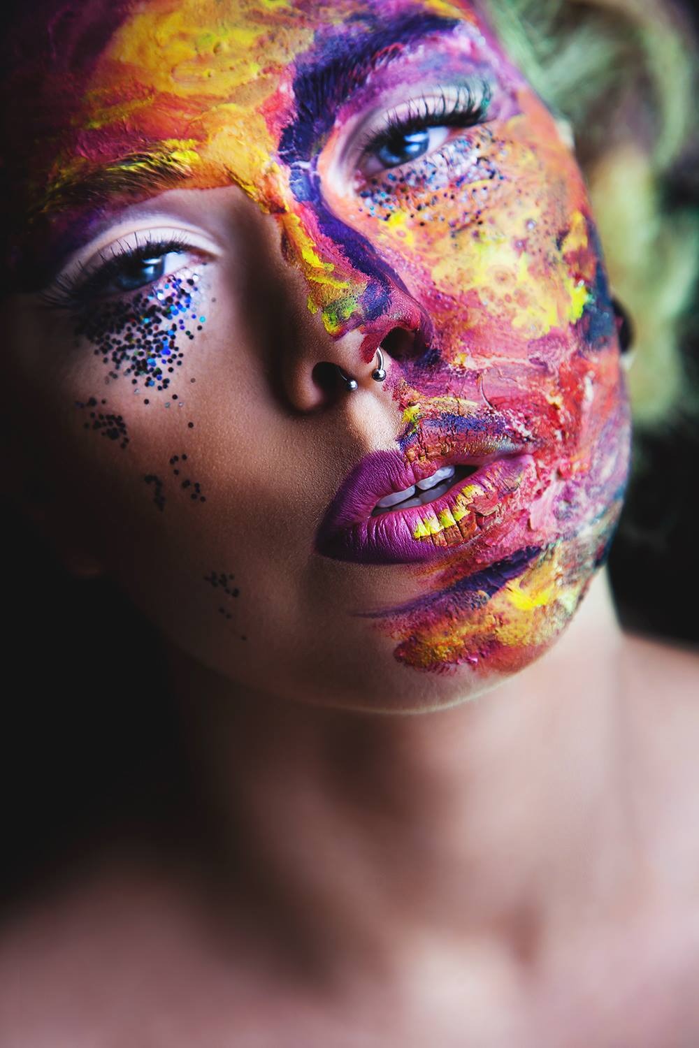 Portret kobiety z artystycznym makijażem twarzy w odcieniach żółci, czerwieni, fioletu i niebieskiego, z brokatem pod oczami i piercingiem w nosie, skierowana w stronę obiektywu.