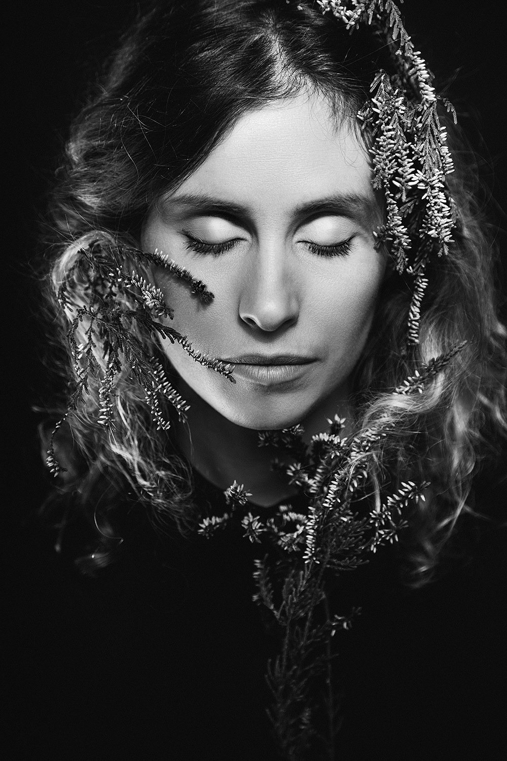 Portret kobiety z zamkniętymi oczami w otoczeniu gałązek wrzosu na czarnym tle, w monochromatycznej tonacji.