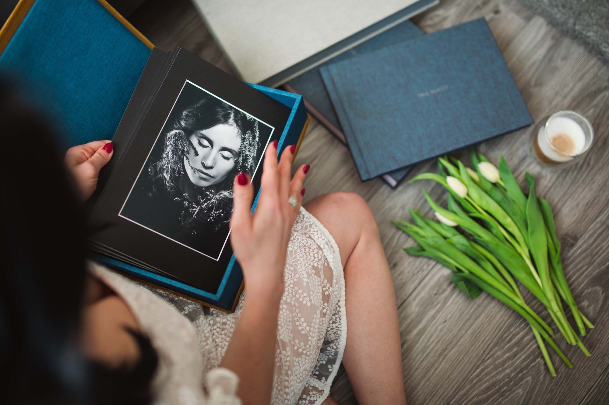 Kobieta w koronkowej sukience przegląda album ze zdjęciami portretowymi w czerni i bieli, obok leżą tulipany i kawa latte.