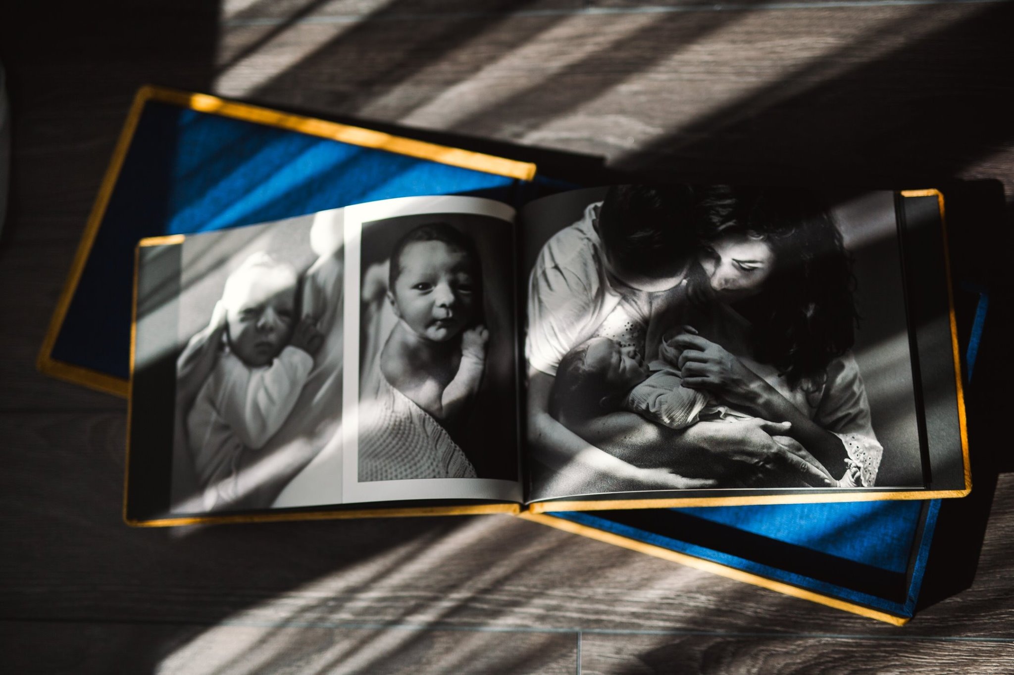 Otwarty album w niebieskiej oprawie ze złotym obramowaniem prezentujący czarno-białe fotografie noworodka, w tym ujęcie matki trzymającej dziecko, oświetlone promieniami słońca.