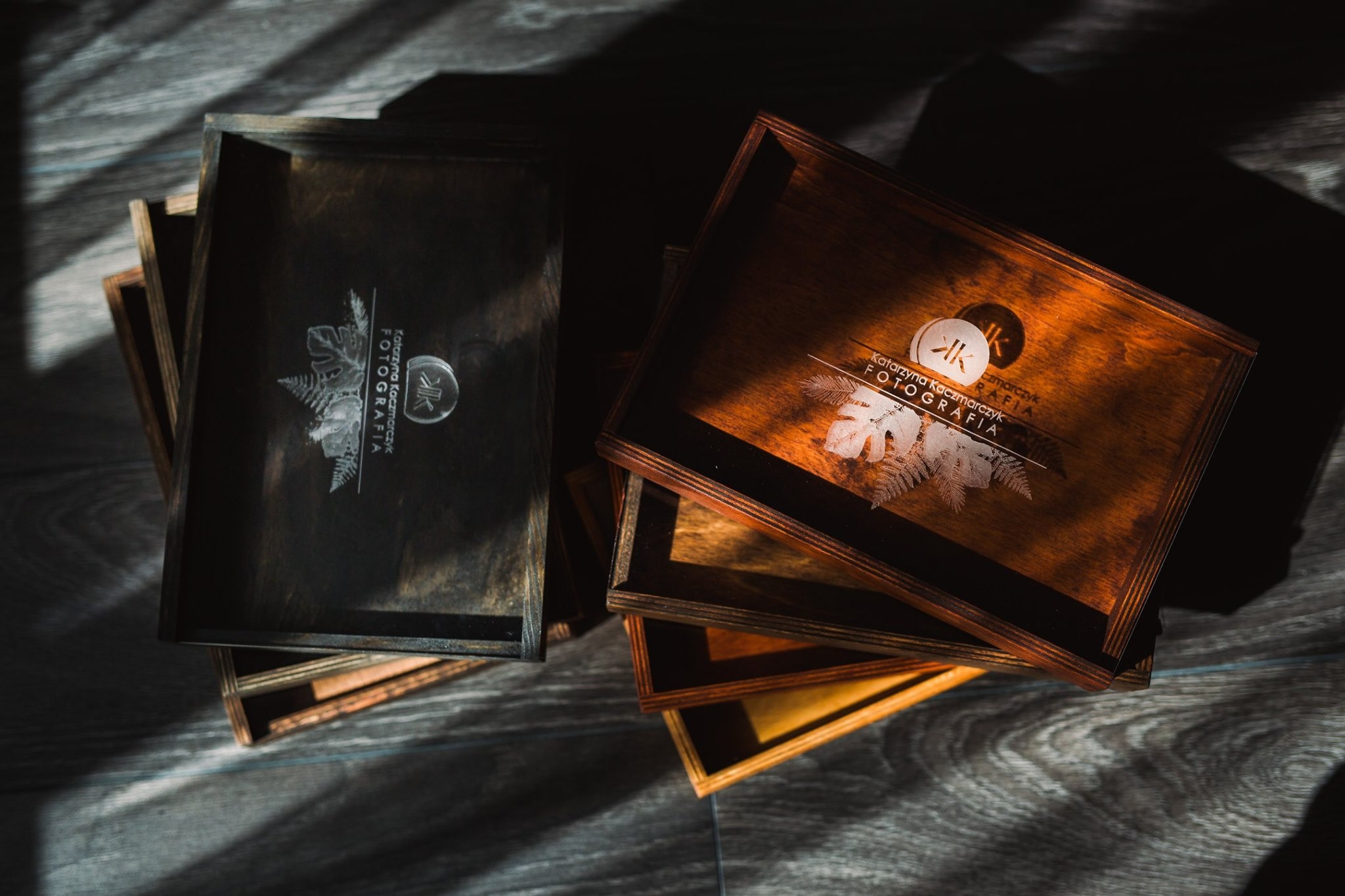 Dwa drewniane pudełka na zdjęcia z wygrawerowanym logo i motywem roślinnym, jedno ciemne, drugie jasne, ułożone na drewnianej powierzchni w promieniach słonecznego światła.