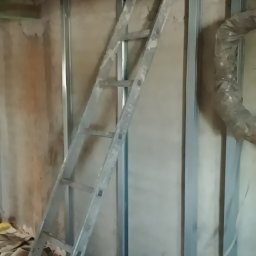 Marcel Szostak - Montaż stelaża z profili stalowych pod płyty gipsowe z widoczną wełną izolacyjną i drabiną opartą o ścianę.