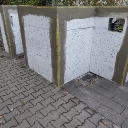 Fragment ocieplonej ściany zewnętrznej budynku z widoczną siatką wzmacniającą i częściowo nałożoną warstwą kleju, przygotowaną do dalszych prac wykończeniowych, z widoczną puszką elektryczną.