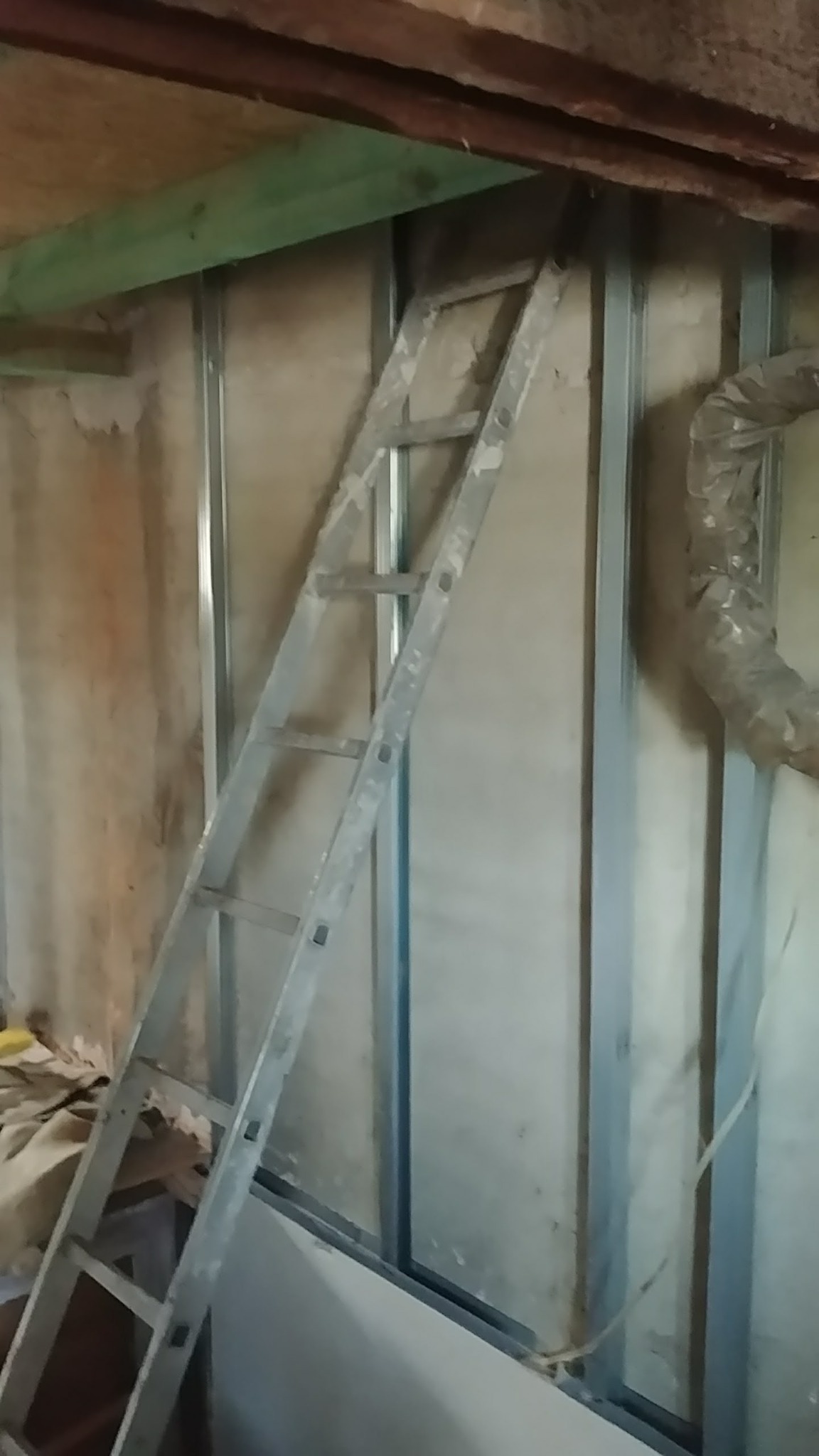 Montaż stelaża z profili stalowych pod płyty gipsowe z widoczną wełną izolacyjną i drabiną opartą o ścianę.