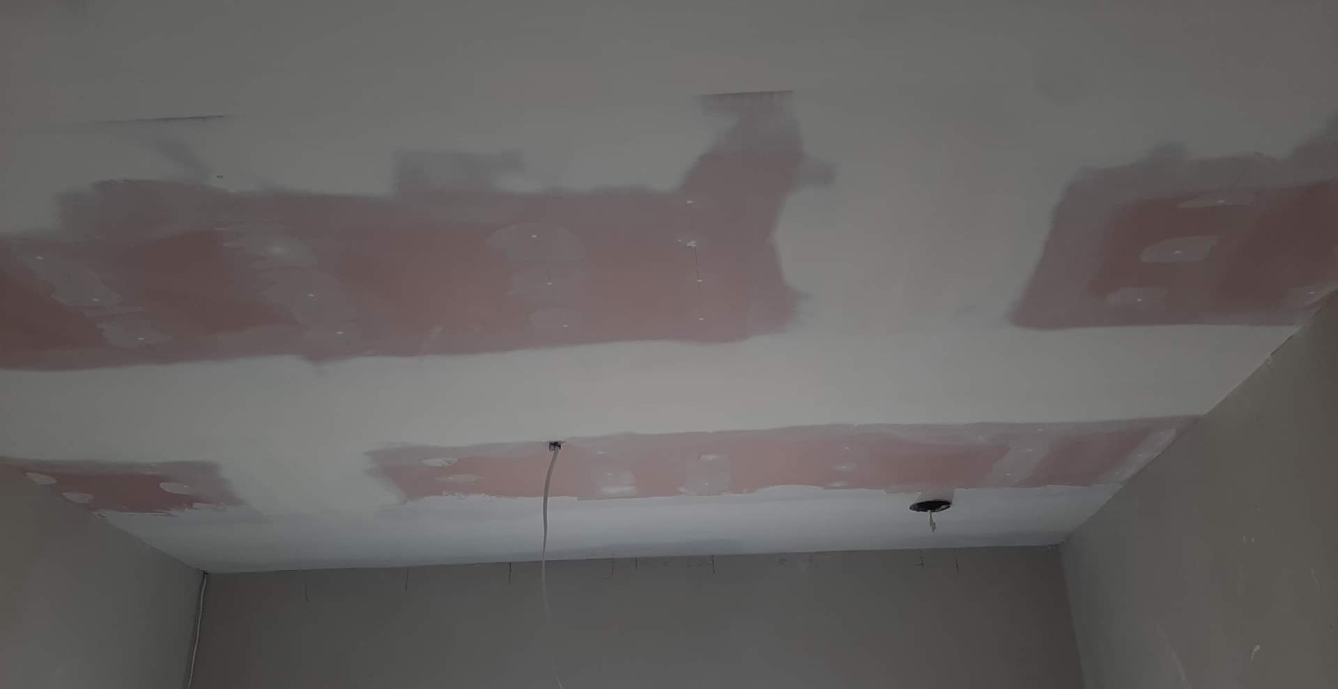 Sufit w trakcie szpachlowania z widocznymi łatami szpachli w kolorze różowym, przygotowywany do malowania, z wystającym przewodem elektrycznym.