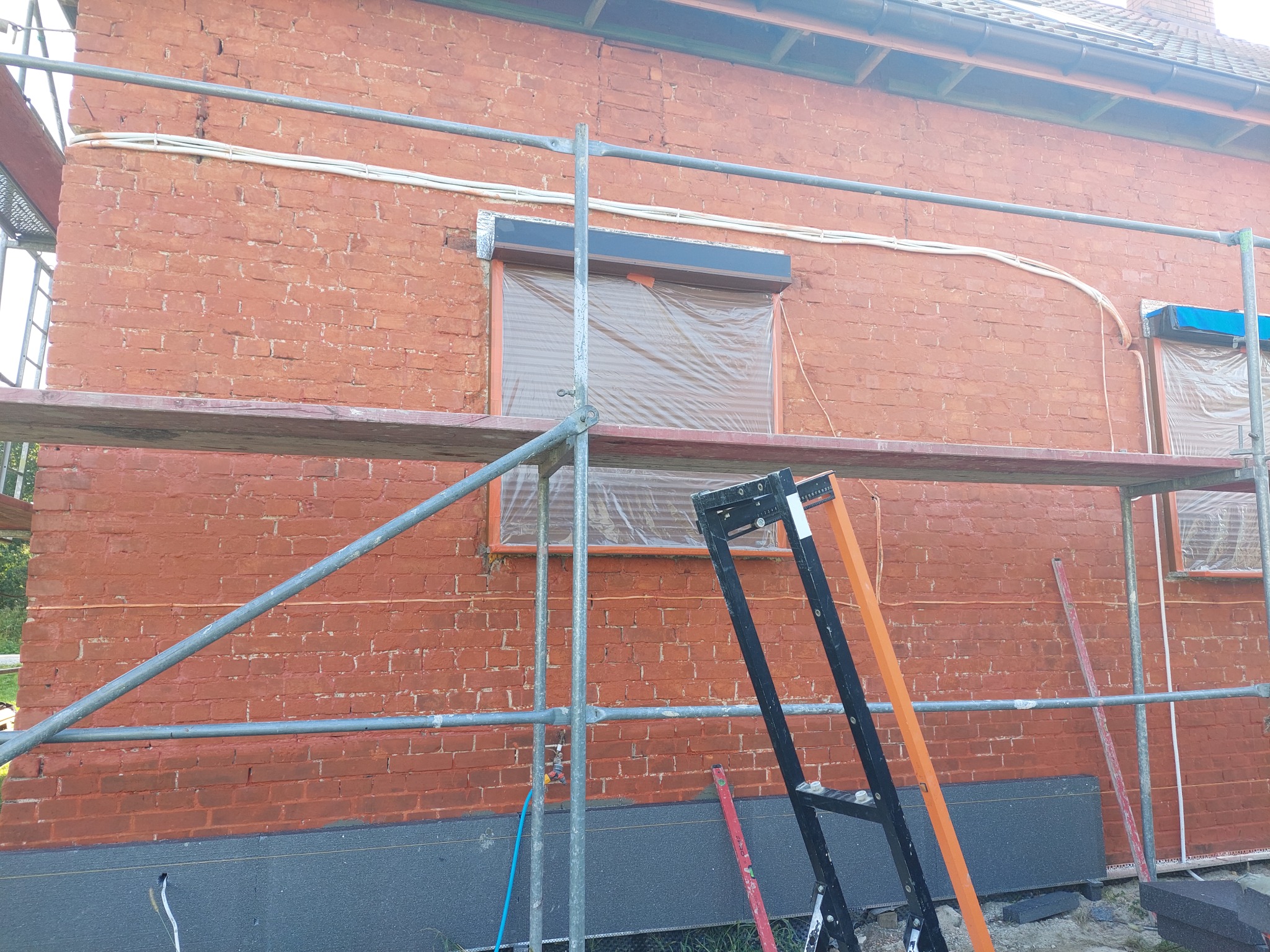 Ceglana ściana budynku w trakcie prac ociepleniowych, widoczne rusztowanie, zabezpieczone okna folią, częściowo ocieplony cokół budynku szarymi płytami styropianowymi.