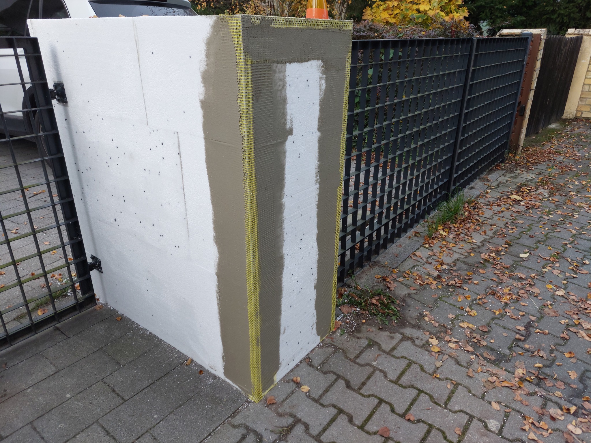 Fragment ocieplonego słupka ogrodzenia z widoczną siatką i warstwą kleju, w tle metalowe ogrodzenie i bruk pokryty liśćmi.