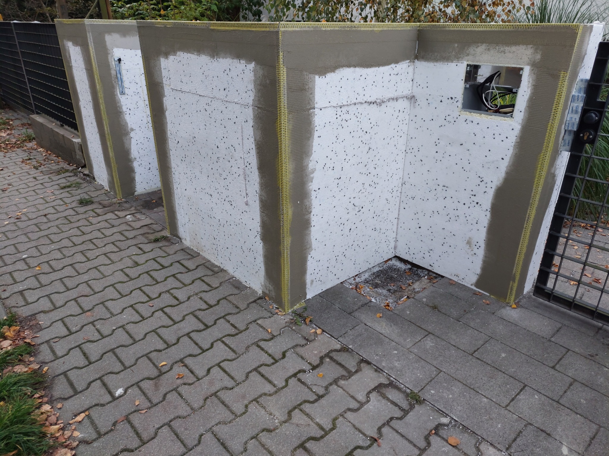 Fragment ocieplonej ściany zewnętrznej budynku z widoczną siatką wzmacniającą i częściowo nałożoną warstwą kleju, przygotowaną do dalszych prac wykończeniowych, z widoczną puszką elektryczną.