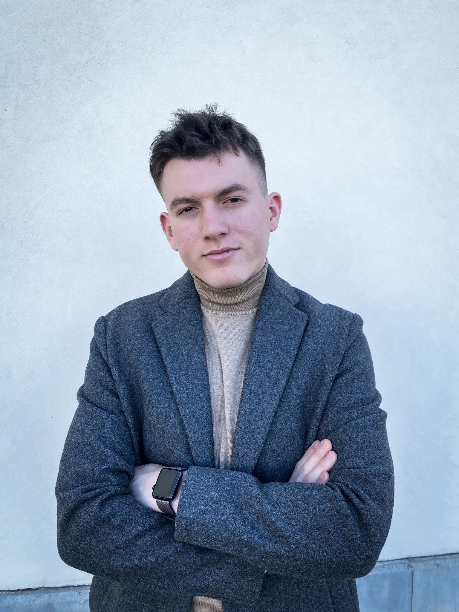 Młody mężczyzna w szarym płaszczu i beżowym golfie pozuje na tle jasnej ściany, z założonymi rękami i widocznym smartwatchem.