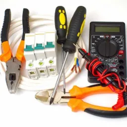 Zestaw narzędzi i akcesoriów elektrycznych na białym tle: multimetr cyfrowy z przewodami pomiarowymi, wyłączniki nadprądowe, przewód elektryczny, kombinerki z pomarańczowymi rękojeściami i śrubokręty.