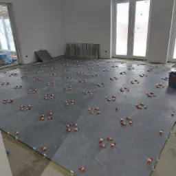 Układanie szarych płytek podłogowych z marmurkowym wzorem, z użyciem klipsów poziomujących, w trakcie prac w Nowym Targu. Widoczne białe ściany i drzwi balkonowe.