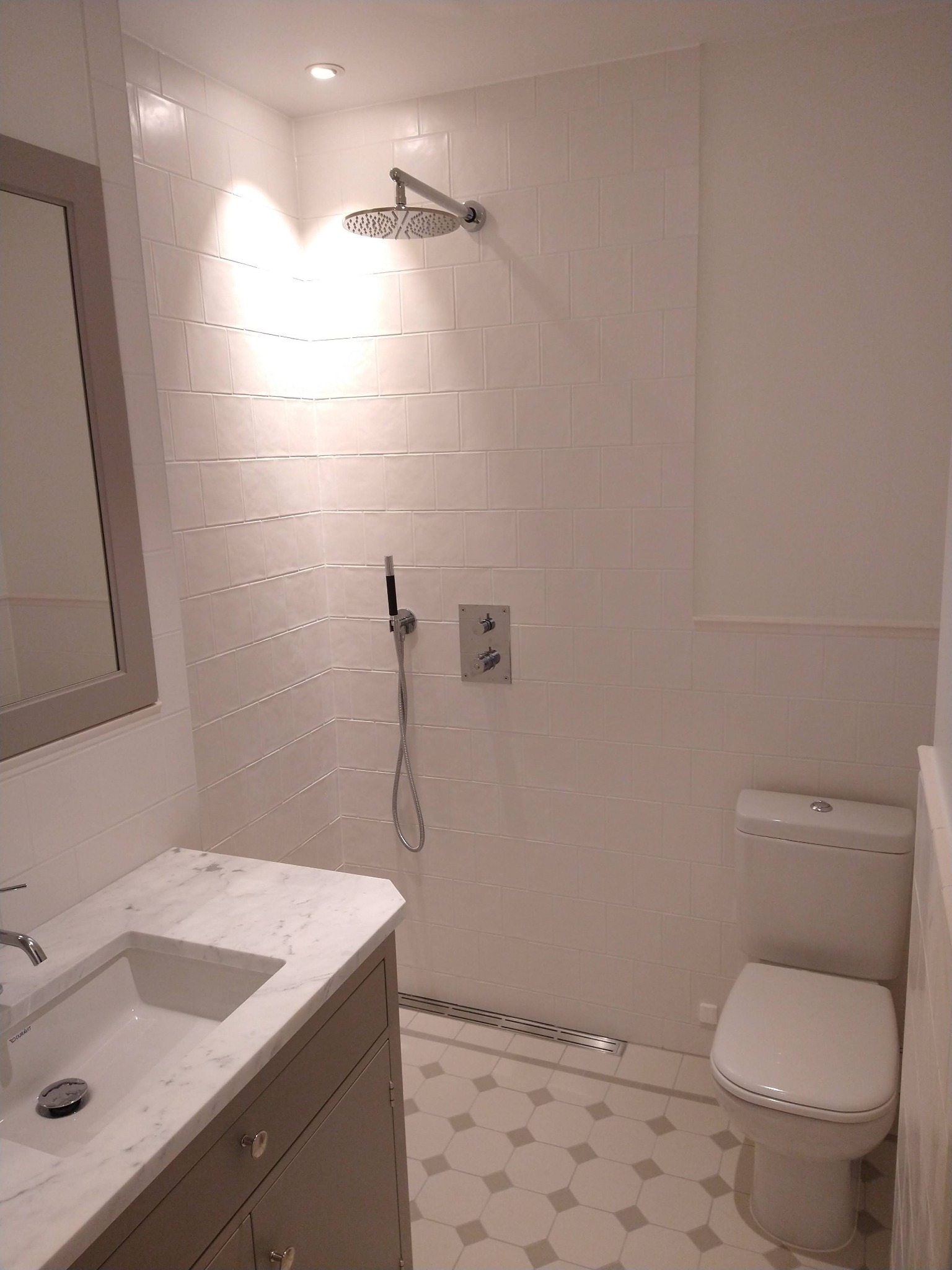 Jasna łazienka z białymi, kwadratowymi płytkami, kabiną prysznicową typu walk-in z deszczownicą, umywalką z marmurowym blatem i toaletą.