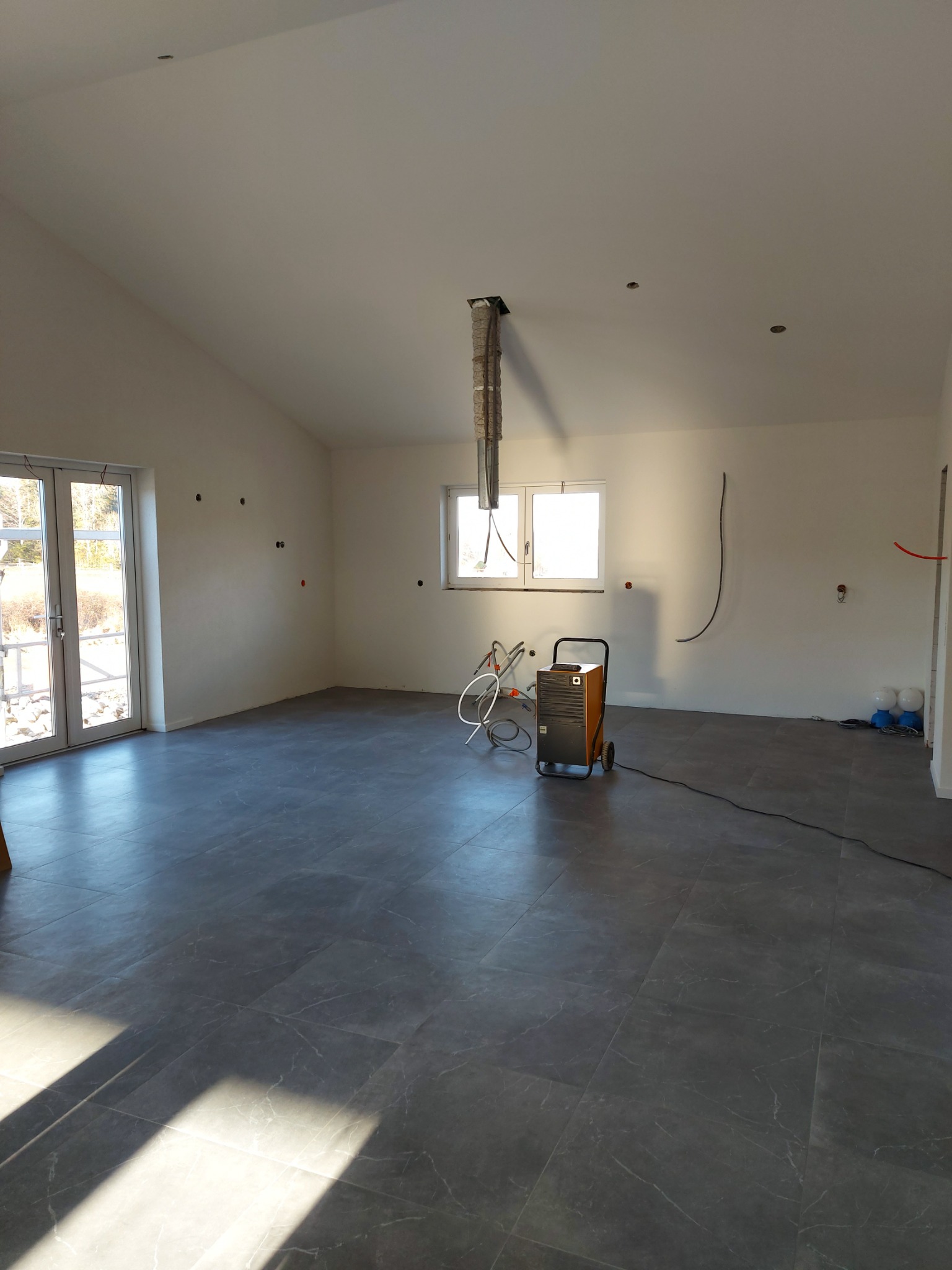 Pomieszczenie w trakcie wykańczania: szare płytki podłogowe, białe ściany z widocznymi punktami elektrycznymi, osuszacz powietrza i kable. Widoczne okno i drzwi balkonowe.