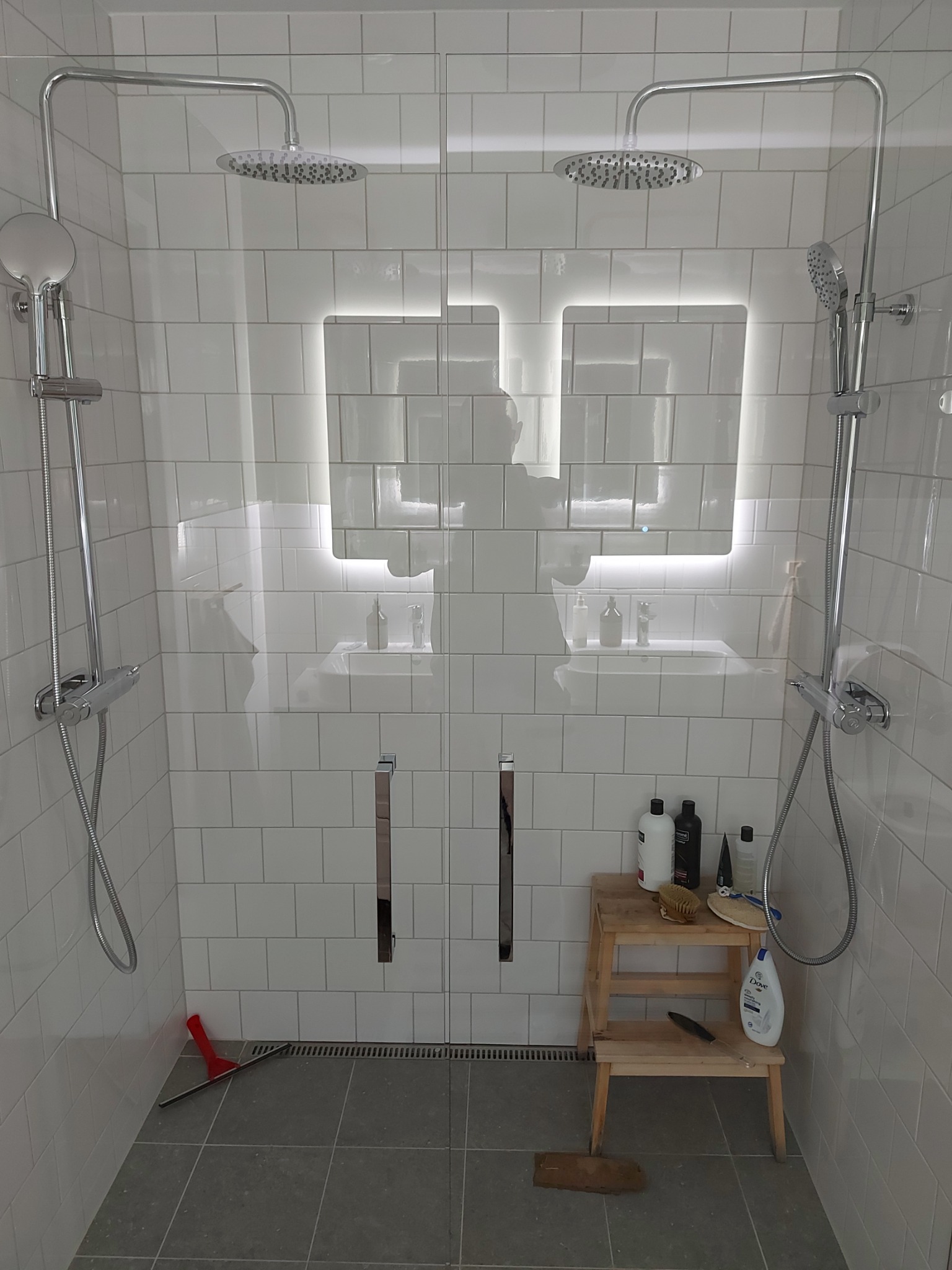 Nowoczesna kabina prysznicowa z dwoma deszczownicami i podświetlanymi lustrami, wykończona białymi, prostokątnymi płytkami, szara podłoga z odpływem liniowym, drewniany stołek z kosmetykami...