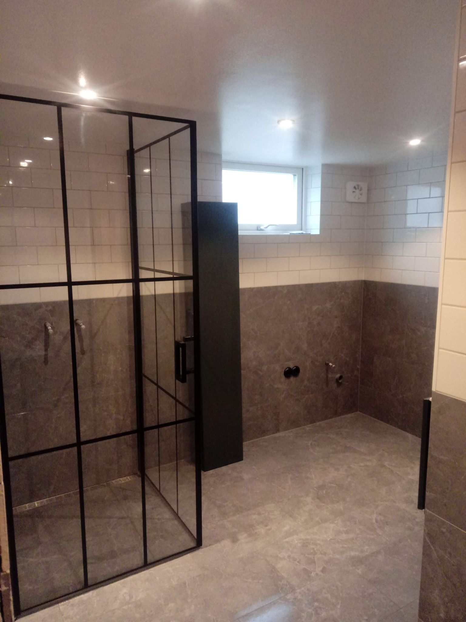 Nowoczesna łazienka z kabiną prysznicową typu walk-in z czarnymi profilami, wykończona płytkami imitującymi marmur w odcieniach szarości i białymi cegiełkami, z widocznymi przyłączami hydraulicznymi.